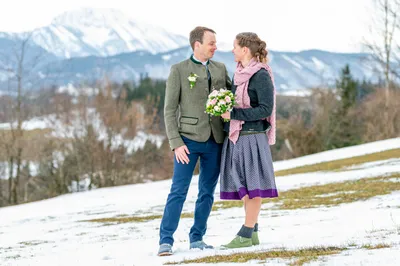 Ein Mann und eine Frau stehen nah beieinander auf einem verschneiten Berg, lächeln und schauen sich an. Der Mann trägt eine grüne Jacke und die Frau hält einen Blumenstrauß.