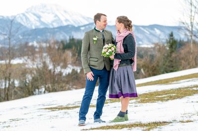 Ein Mann und eine Frau stehen nah beieinander auf einem verschneiten Berg, lächeln und schauen sich an. Der Mann trägt eine grüne Jacke und die Frau hält einen Blumenstrauß.