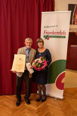 Ein älterer Mann und eine Frau stehen vor einem Banner, der Mann hält eine Holztafel mit einer Uhr und die Frau hält einen Blumenstrauß.
