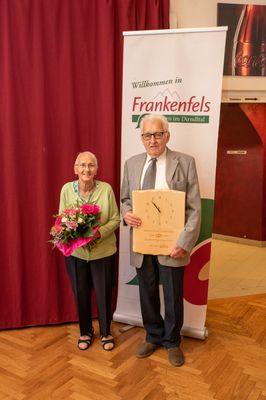 Ein älteres Paar steht vor einem Banner, das 'Willkommen in Frankenfels im Dirndital' besagt. Der Mann hält eine Holzuhr und die Frau einen Blumenstrauß.