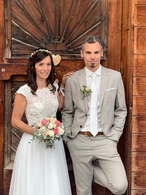 Eine lächelnde Braut in einem weißen Spitzenkleid und ein Bräutigam in einem hellgrauen Anzug stehen gemeinsam vor einer Holztür.