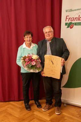 Ein älterer Mann und eine ältere Frau stehen zusammen und halten eine Holztafel und einen Blumenstrauß. Der Mann trägt eine Brille und eine Jacke, während die Frau einen hellblauen Pullover trägt. Hinter ihnen ist ein roter Vorhang und ein grünes Banner mit einem Logo.