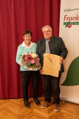 Ein älterer Mann und eine ältere Frau stehen zusammen und halten eine Holztafel und einen Blumenstrauß. Der Mann trägt eine Brille und eine Jacke, während die Frau einen hellblauen Pullover trägt. Hinter ihnen ist ein roter Vorhang und ein grünes Banner mit einem Logo.