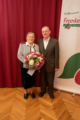 Ein älteres Paar steht lächelnd zusammen mit einem Blumenstrauß vor sich, vor einem roten Hintergrund mit einem grünen Blattlogo.
