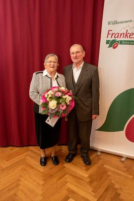 Ein älteres Paar steht lächelnd zusammen mit einem Blumenstrauß vor sich, vor einem roten Hintergrund mit einem grünen Blattlogo.