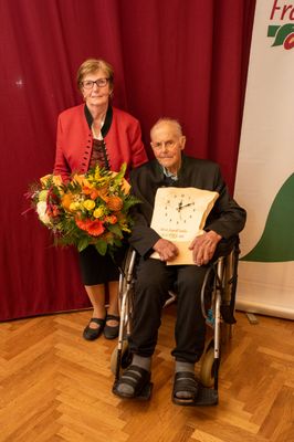 Ein älterer Mann im Rollstuhl hält eine Holzuhr und einen Blumenstrauß, während eine Frau neben ihm vor einem roten Vorhang steht.