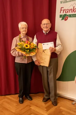 Ein älteres Paar steht lächelnd nebeneinander, die Frau hält einen Blumenstrauß und der Mann eine Holzuhr und ein Zertifikat. Sie stehen vor einem roten Vorhang.