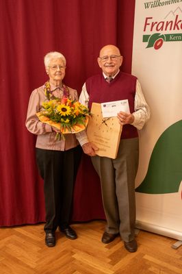 Ein älteres Paar steht lächelnd nebeneinander, die Frau hält einen Blumenstrauß und der Mann eine Holzuhr und ein Zertifikat. Sie stehen vor einem roten Vorhang.