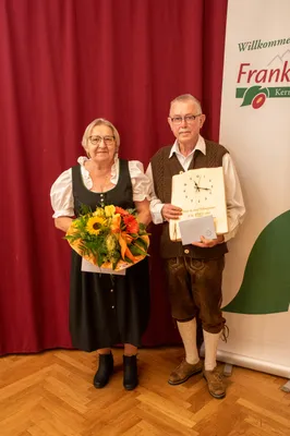 Ein älteres Paar steht vor einem roten Vorhang, die Frau hält einen Blumenstrauß und der Mann eine gerahmte Uhr und ein Zertifikat.
