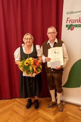 Ein älteres Paar steht vor einem roten Vorhang, die Frau hält einen Blumenstrauß und der Mann eine gerahmte Uhr und ein Zertifikat.