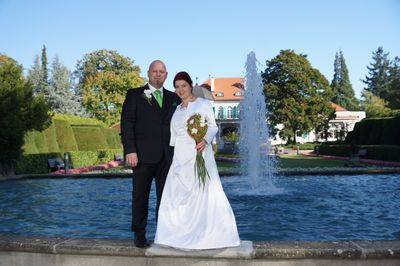 Ein frisch verheiratetes Paar steht vor einem Brunnen, die Braut hält einen Blumenstrauß in der Hand und der Bräutigam trägt eine grüne Krawatte. Hinter ihnen steht ein Gebäude mit orangefarbenen Dächern und Bäumen.