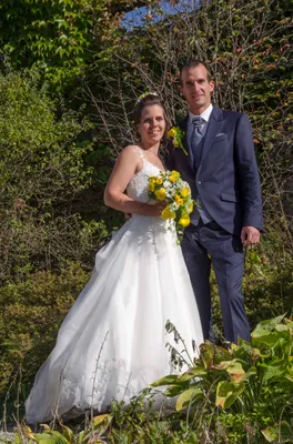 Ein frisch verheiratetes Paar steht eng beieinander, die Braut in einem weißen Kleid und der Bräutigam in einem dunklen Anzug, beide lächeln und halten einen Blumenstrauß.