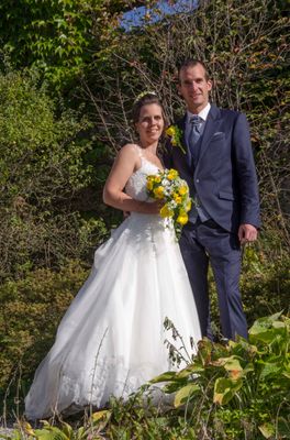 Ein frisch verheiratetes Paar steht eng beieinander, die Braut in einem weißen Kleid und der Bräutigam in einem dunklen Anzug, beide lächeln und halten einen Blumenstrauß.