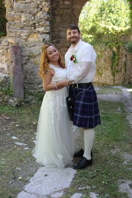 Ein frisch verheiratetes Paar steht draußen, die Frau in einem weißen Hochzeitskleid und der Mann in einem Kilt, lächelnd für die Kamera.