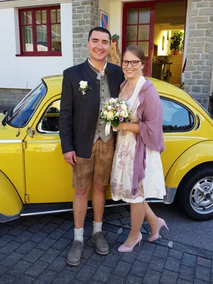 Ein Mann und eine Frau stehen neben einem gelben Auto, lächeln und halten einen Blumenstrauß. Der Mann trägt ein traditionelles Outfit und die Frau ein weißes Kleid mit einem rosa Schal.