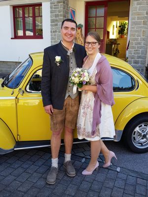 Ein Mann und eine Frau stehen neben einem gelben Auto, lächeln und halten einen Blumenstrauß. Der Mann trägt ein traditionelles Outfit und die Frau ein weißes Kleid mit einem rosa Schal.
