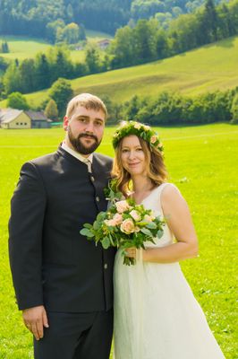 Ein frisch verheiratetes Paar steht auf einem üppigen grünen Feld, der Mann in einem schwarzen Anzug und die Frau in einem weißen Kleid mit Blumenkranz.