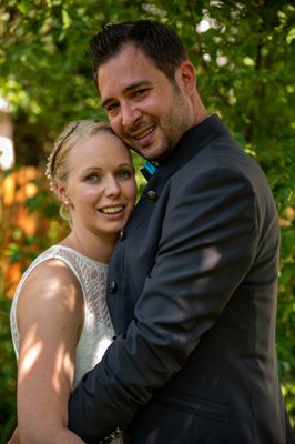 Ein lächelndes Paar steht eng zusammen in einem Garten, der Mann in einem schwarzen Anzug und die Frau in einem weißen Kleid.