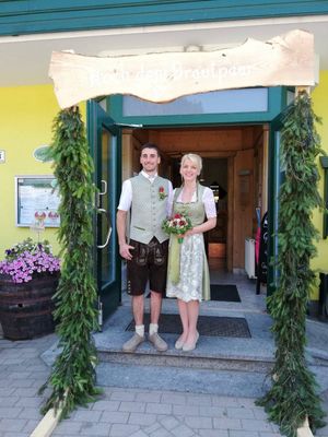 Ein lächelndes Paar steht vor einem geschmückten Eingang, der Mann in traditioneller Kleidung und die Frau mit einem Blumenstrauß. Hinter ihnen ist eine gelbe Wand mit einem Schild und einem Fass mit Blumen.