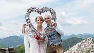 Ein frisch verheiratetes Paar steht auf einem Berggipfel, hält einen herzförmigen Rahmen aus Zweigen und lächelt in die Kamera.