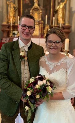 Ein frisch verheiratetes Paar steht nah beieinander, lächelt, der Bräutigam trägt einen grünen Mantel und die Braut ein weißes Hochzeitskleid, hält einen Blumenstrauß.