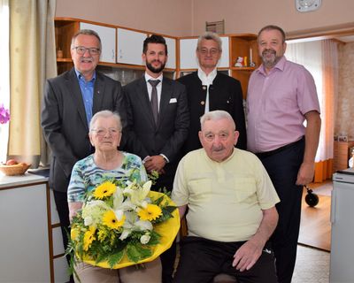 Eine Gruppe von Menschen, darunter ein älteres Paar, steht in einem Raum mit einem Blumenstrauß im Vordergrund.