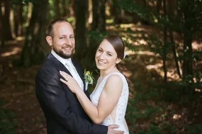 Ein frisch verheiratetes Paar steht eng beieinander in einem Wald, der Bräutigam in einem schwarzen Anzug und die Braut in einem weißen Hochzeitskleid, beide lächeln für die Kamera.