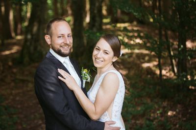 Ein frisch verheiratetes Paar steht eng beieinander in einem Wald, der Bräutigam in einem schwarzen Anzug und die Braut in einem weißen Hochzeitskleid, beide lächeln für die Kamera.