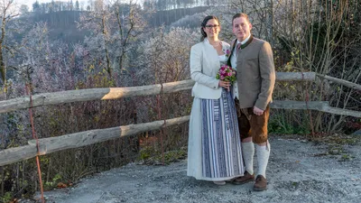 Ein frisch verheiratetes Paar steht eng beieinander, lächelt, die Frau hält einen Blumenstrauß, beide in traditioneller Kleidung, vor einem Hintergrund von frostbedeckten Bäumen.