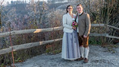 Ein frisch verheiratetes Paar steht eng beieinander, lächelt, die Frau hält einen Blumenstrauß, beide in traditioneller Kleidung, vor einem Hintergrund von frostbedeckten Bäumen.