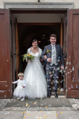 Ein frisch verheiratetes Paar und ein kleines Mädchen stehen vor einer Kirche, umgeben von Seifenblasen. Die Braut trägt ein weißes Kleid und der Bräutigam einen grauen Anzug. Das Mädchen ist in ein weißes Kleid mit einer Blumenkrone gekleidet.