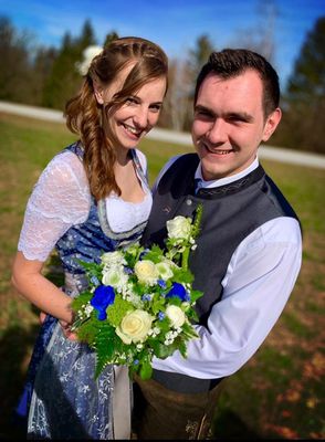 Ein lächelndes Paar in traditioneller Kleidung, der Mann hält einen Strauß weißer und blauer Blumen, steht auf einer Wiese.