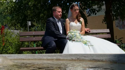 Ein frisch verheiratetes Paar, der Bräutigam im Anzug und die Braut im Hochzeitskleid, sitzt auf einer Bank mit einem Brunnen im Vordergrund.