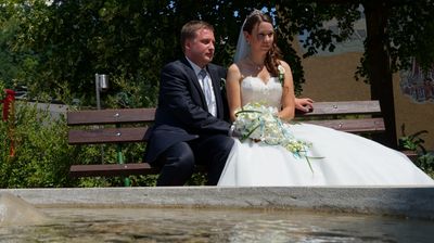 Ein frisch verheiratetes Paar, der Bräutigam im Anzug und die Braut im Hochzeitskleid, sitzt auf einer Bank mit einem Brunnen im Vordergrund.