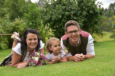 Eine lächelnde Familie mit drei Personen liegt auf einem grünen Rasen, wobei die Frau einen Blumenstrauß hält, der Mann eine Brille trägt und ein kleines Mädchen in der Mitte lächelt.