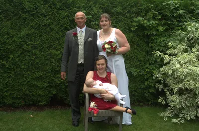 Eine vierköpfige Familie steht in einem Garten, mit einem Mann und einer Frau auf beiden Seiten einer Frau, die ein Baby hält. Die Frau hält einen Blumenstrauß.