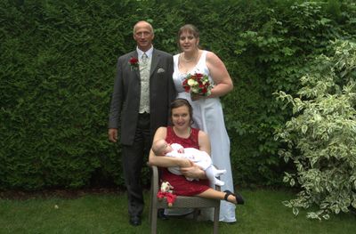 Eine vierköpfige Familie steht in einem Garten, mit einem Mann und einer Frau auf beiden Seiten einer Frau, die ein Baby hält. Die Frau hält einen Blumenstrauß.