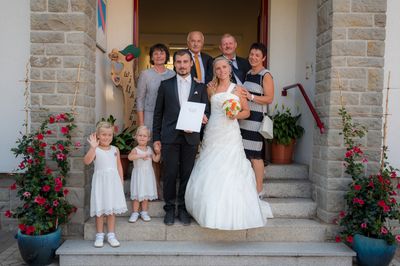 Ein frisch verheiratetes Paar steht mit Familienmitgliedern auf den Stufen eines Gebäudes, hält ein Zertifikat und einen Blumenstrauß. Zwei junge Mädchen winken im Vordergrund.