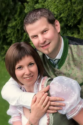 Ein lächelndes Paar in traditioneller deutscher Kleidung, der Mann hält ein Baby in einem Blumenkleid, steht in einem grünen Garten.
