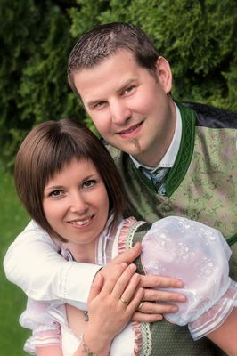 Ein lächelndes Paar in traditioneller deutscher Kleidung, der Mann hält ein Baby in einem Blumenkleid, steht in einem grünen Garten.