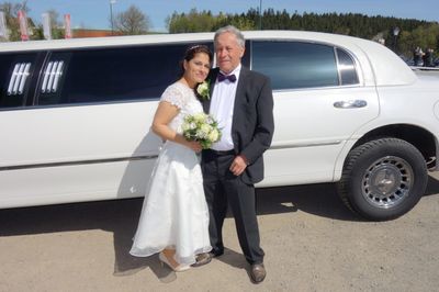 Ein frisch verheiratetes Paar steht vor einer weißen Limousine, der Bräutigam trägt eine Fliege und die Braut hält einen Blumenstrauß.