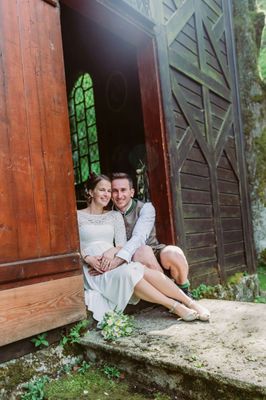 Ein lächelndes Paar, die Braut in einem weißen Kleid und der Bräutigam in einem Weste, sitzt vor einer Holztür mit einem grünen Fenster dahinter.