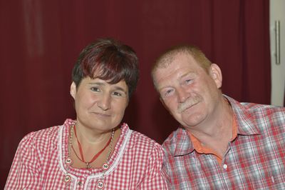 Ein Mann und eine Frau lächeln und posieren für ein Foto. Die Frau trägt ein rot-weiß kariertes Oberteil und eine Halskette mit roten Anhängern. Der Mann trägt ein rot-grau kariertes Hemd. Sie stehen vor einem roten Vorhang.