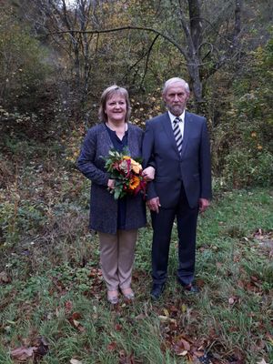 Ein älteres Paar steht in einem Feld mit Herbstlaub, die Frau hält einen Blumenstrauß, beide lächeln.