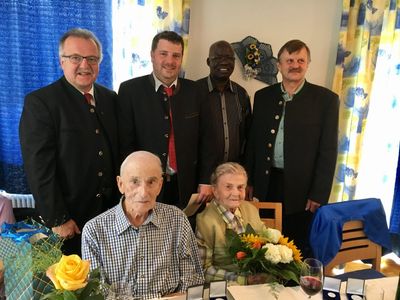 Eine Gruppe von fünf Personen, zwei Männer und drei Frauen, stehen und sitzen vor einem Tisch mit Blumen und Weingläsern. Sie lächeln und scheinen für ein Foto zu posieren.