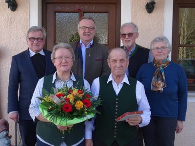 Eine Gruppe älterer Menschen steht vor einer Tür, einer hält einen Blumenstrauß, ein anderer ein Buch und andere lächeln.