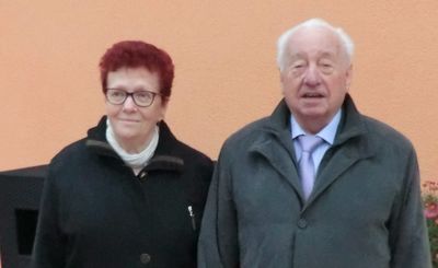 Ein älterer Mann und eine ältere Frau stehen nebeneinander und lächeln. Die Frau trägt eine Brille und einen weißen Schal, während der Mann eine lila Krawatte trägt. Sie stehen vor einer pfirsichfarbenen Wand.