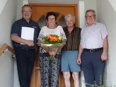Vier Personen stehen vor einer Holztür, eine hält einen Blumenstrauß. Die Frau in der Mitte trägt eine Brille und eine Halskette. Der Mann links hält ein Papier.