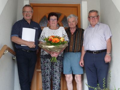 Vier Personen stehen vor einer Holztür, eine hält einen Blumenstrauß. Die Frau in der Mitte trägt eine Brille und eine Halskette. Der Mann links hält ein Papier.