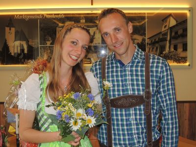 Ein lächelndes Paar in traditioneller deutscher Kleidung, die Frau hält einen Blumenstrauß, steht vor einem Hintergrund mit einer Dorfszene.